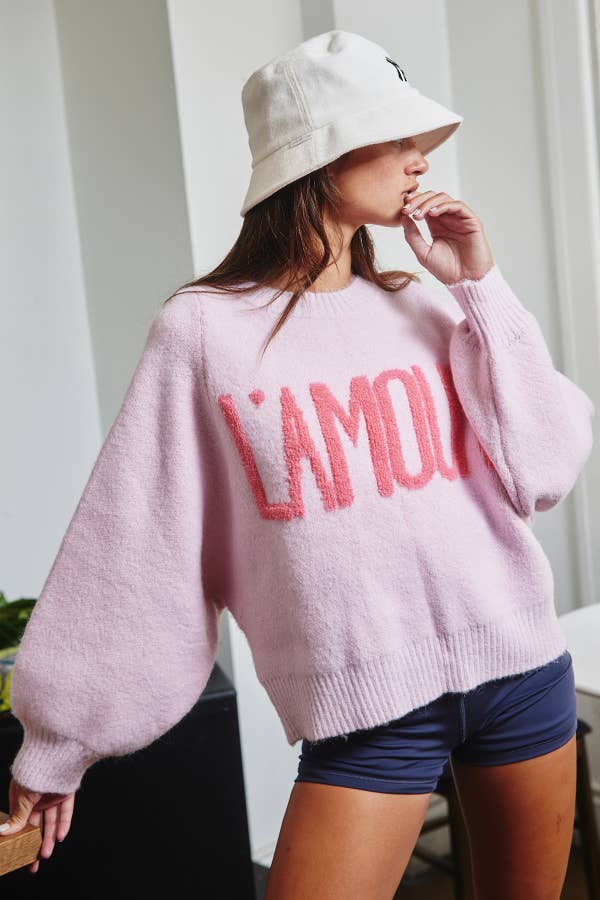 Person wearing a pink sweater with 'L'Amour' text and a white bucket hat indoors.